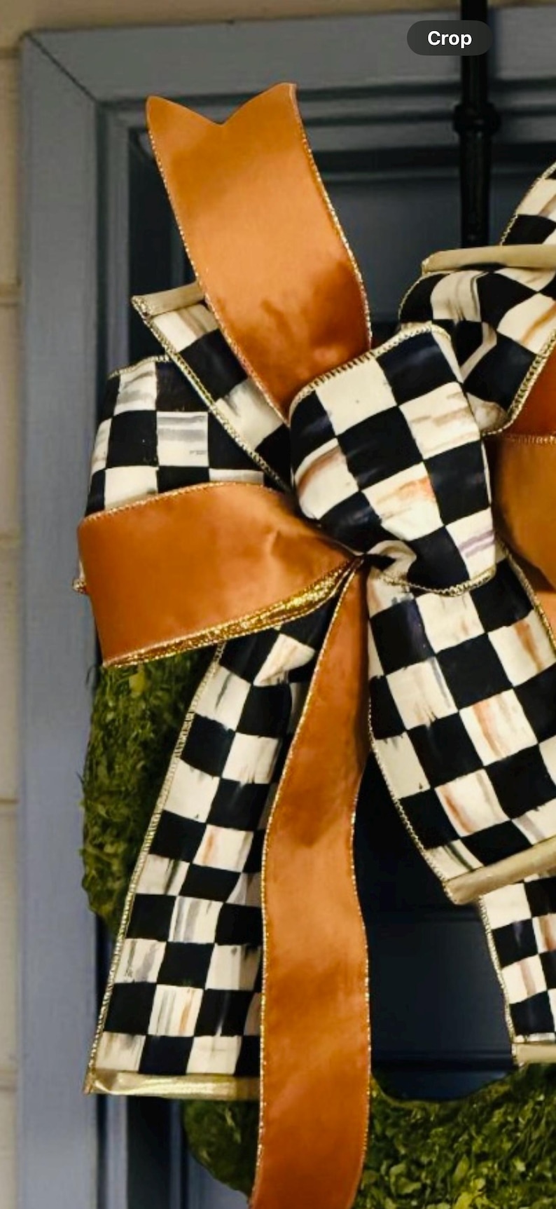 May include: A decorative bow featuring a combination of solid copper-colored ribbon and a black and white checkered pattern ribbon. The bow has multiple loops and long trailing ends with gold-toned edges, attached to a green textured base.
