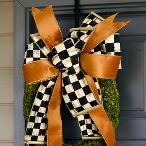 May include: A decorative wreath featuring a large, multi-layered bow made of solid copper-colored ribbon and black and white checkered ribbon. The bow is attached to a circular base covered in dense, textured green foliage, creating a full appearance.