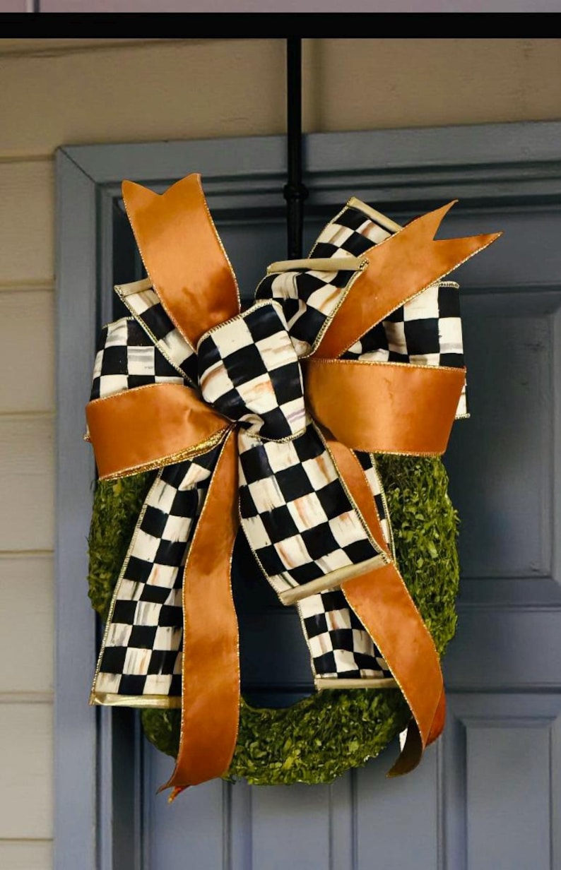 May include: A decorative wreath featuring a large, multi-layered bow made of solid copper-colored ribbon and black and white checkered ribbon. The bow is attached to a circular base covered in dense, textured green foliage, creating a full appearance.