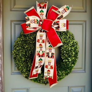 May include: A Christmas wreath with a bright red bow, decorated with a nutcracker pattern. The bow has gold edging and is set against a dense, green, circular wreath, ideal for festive home decoration.