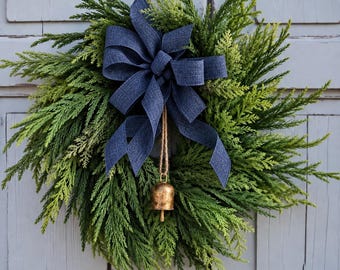 May include: A circular wreath made of lush green cedar foliage featuring a large, textured navy blue fabric bow at the top center. A single metallic gold-toned bell hangs from the center of the bow by a thin, natural-colored jute twine cord.