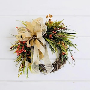 May include: A fall-themed wreath with a natural grapevine base. It features green foliage, red berries, and a large, tan burlap bow. The wreath is decorated with small brown accents and is set against a white background.