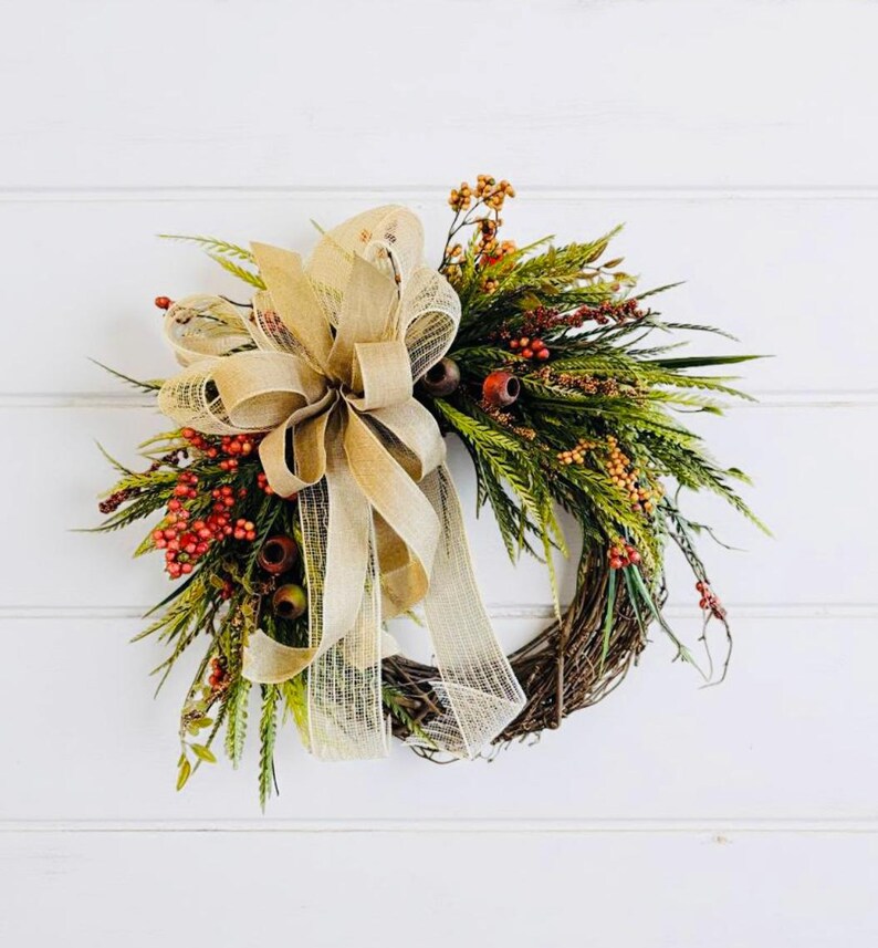 May include: A fall-themed wreath with a natural grapevine base. It features green foliage, red berries, and a large, tan burlap bow. The wreath is decorated with small brown accents and is set against a white background.