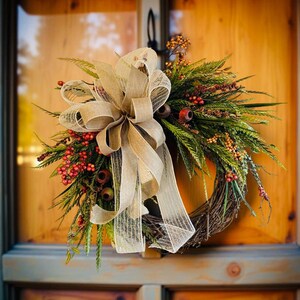 May include: A fall-themed wreath with a woven grapevine base. It features green foliage, red berries, and small orange accents. A large, beige burlap bow is centered on the wreath, adding a rustic touch. The wreath is displayed on a wooden door.