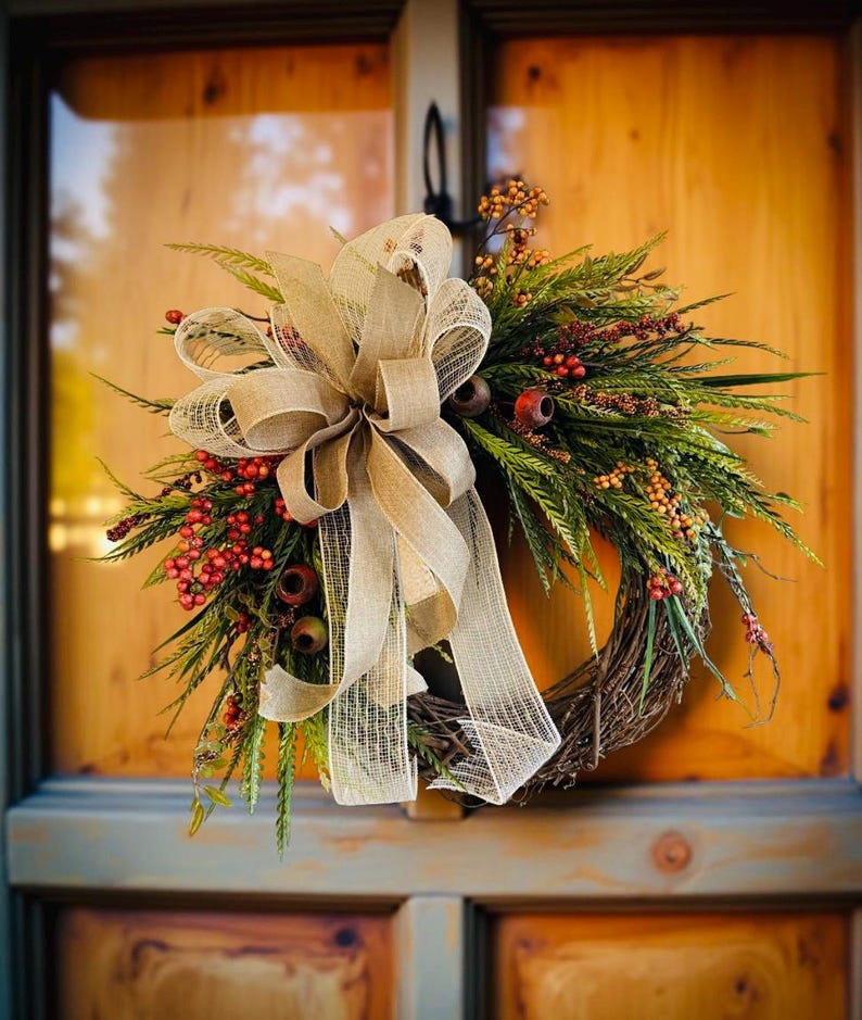 May include: A fall-themed wreath with a woven grapevine base. It features green foliage, red berries, and small orange accents. A large, beige burlap bow is centered on the wreath, adding a rustic touch. The wreath is displayed on a wooden door.