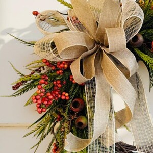 May include: A decorative wreath featuring a large burlap bow and accents of red berries, green foliage, and small brown accents. The wreath is designed for seasonal home decor.