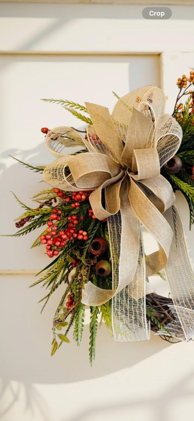 May include: A decorative wreath featuring a large burlap bow and accents of red berries, green foliage, and small brown accents. The wreath is designed for seasonal home decor.