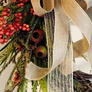May include: A rustic wreath featuring a large burlap bow with a woven texture. The wreath is adorned with red berries, green foliage, and decorative elements, creating a natural and inviting aesthetic.