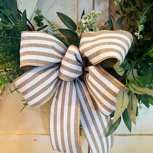 May include: A close-up of a decorative wreath with a large bow made from brown and white striped ribbon. The wreath is decorated with green foliage and small white flowers, giving it a natural appearance.