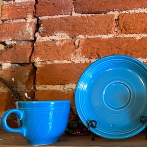 May include: Three turquoise blue ceramic teacups and a saucer. The cups and saucer have a glossy finish. The cups have a rounded shape and a handle. The saucer has a slightly raised rim. The cups and saucer are sitting on a wooden surface in front of a brick wall.