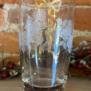 May include: A clear glass tumbler with a floral design and a gold bow. The tumbler features white roses and gold ribbon accents. The glass is cylindrical and appears to be vintage. The background is a brick wall.