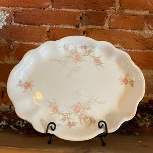 Puede incluir: Una fuente ovalada de color blanco roto con borde festoneado. La fuente está decorada con motivos florales rosas y verdes. Se apoya en un soporte de metal negro, mostrando una estética vintage.