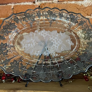 Puede incluir: Un plato de servir de vidrio transparente con un diseño de vid de color plateado. El plato tiene forma ovalada y un borde festoneado. Está sobre un soporte de metal negro.