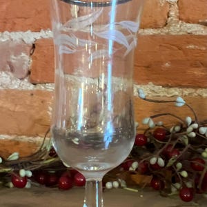 Puede incluir: Un jarrón de vidrio transparente con un borde plateado y un diseño floral blanco grabado. El jarrón tiene una forma alta y esbelta con una base redonda.