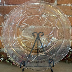 Puede incluir: Un plato de vidrio transparente con un diseño floral en relieve ornamentado. El plato es redondo con un borde festoneado y se exhibe sobre un soporte de metal negro. El plato es probablemente vintage o antiguo.