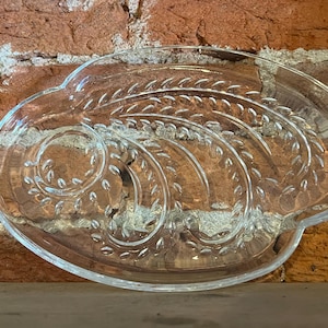 May include: Clear glass serving dish with an oval shape and a decorative leaf design. The dish has a slightly raised edge and is set against a brick wall background. The leaf design is etched into the glass.