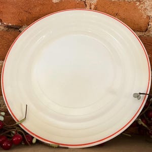 May include: A white ceramic plate with a red rim. The plate has a simple, classic design with concentric rings. The plate is set against a brick background, suggesting a rustic or farmhouse aesthetic. This plate is suitable for serving food.