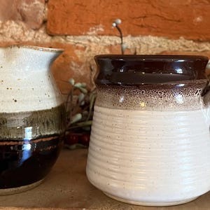May include: Two ceramic mugs. The mug on the left is a small pitcher with a ruffled top, white, brown, and black glaze. The mug on the right is a white mug with a brown rim and handle. The mugs are on a wooden surface.