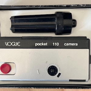 May include: A silver and black Vogue pocket 110 camera with a red shutter button and a separate black flash attachment, presented in a black box with a white molded plastic insert. The camera body features the text Vogue pocket 110 camera.