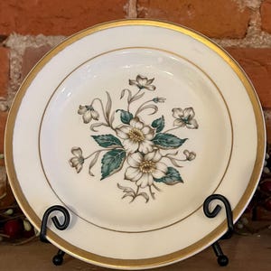 May include: A vintage cream-colored plate with a gold rim and floral design. The center features a cluster of white flowers with brown centers and green leaves. The plate is displayed on a black metal stand.