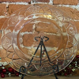May include: A clear glass plate with a pink tint and a raised floral design. The plate has a scalloped edge and is on a black stand.