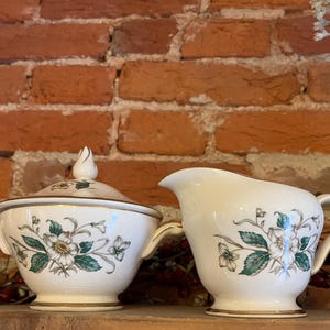 Puede incluir: Azucarera color crema con tapa y jarra de crema a juego. Ambas piezas presentan un diseño floral con flores blancas, hojas verdes y ribetes dorados. La azucarera tiene asas y un adorno decorativo en la tapa. La jarra de crema tiene asa y boquilla.