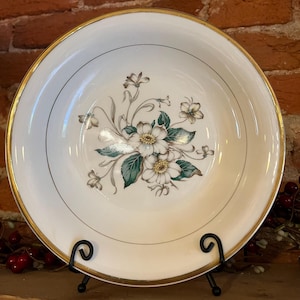 May include: A white ceramic bowl with a gold rim and a floral design. The design features white flowers with yellow centers, green leaves, and gray accents. The bowl is displayed on a black metal stand.