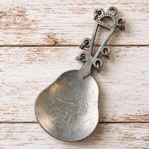 May include: A silver-toned metal spoon featuring a pear-shaped bowl with an engraved deer illustration. The handle is elongated and triangular with a series of small circular loops along the sides and a small ring at the top for hanging.