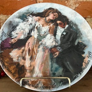 May include: A decorative plate featuring a romantic painting of a couple in period attire. The artwork depicts a woman in a white dress and a man in a black suit. The plate has text in French and the words "Sarah Bernhardt" are visible.