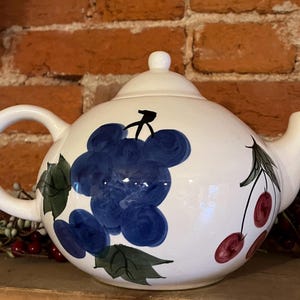 May include: White ceramic teapot with a round body, a curved handle, and a spout. The teapot is decorated with hand-painted blue grapes and red cherries with green leaves. The lid has a small white knob.