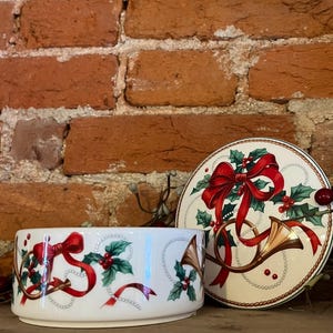 May include: A white ceramic bowl and lid with a Christmas theme. The bowl and lid feature red ribbons, holly, and gold-colored horns. The bowl is round with a low profile.