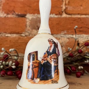 May include: White ceramic bell with a white handle and gold trim. The bell features a painted illustration of a woman seated, reading a letter. The artwork is signed "Norman Rockwell".
