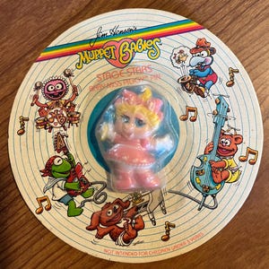 May include: A round Jim Henson's Muppet Babies Stage Stars Baby Miss Piggy pin on a circular cardboard backing. The backing features colorful illustrations of Muppet characters playing musical instruments and musical notes, with a rainbow stripe.