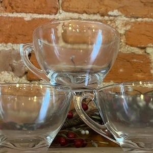 Könnte beinhalten: Drei transparente Teetassen mit Henkeln sind pyramidenförmig gestapelt. Die transparenten Tassen lassen Licht durch. Der Hintergrund ist eine rustikale Backsteinmauer, und die Tassen stehen auf einem dekorativen Sockel.