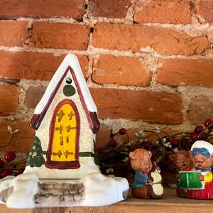 May include: A ceramic Christmas village house with a yellow door and snow-covered roof, flanked by two bear figurines. The house has a green tree and a stone-like base. The bears are singing carols.