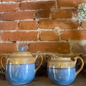 May include: A vintage blue and gold ceramic sugar bowl with a lid and a matching creamer. The sugar bowl has a gold handle and a blue base. The creamer has a gold handle and a blue base. The background is a brick wall.