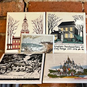 Puede incluir: Una colección de azulejos de cerámica vintage con varios monumentos y escenas. Los azulejos representan edificios, paisajes y texto, incluyendo "Atlantic City Souvenir", "Washington's Headquarters" y "Sandwich Glass Museum". Los azulejos están dispuestos sobre una superficie de madera.