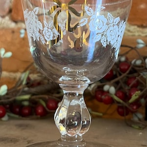 May include: A clear glass goblet with a floral and bow design. The goblet features a round base, a decorative stem, and a bowl-shaped top. White floral patterns and a gold bow are etched on the glass, adding a touch of elegance. The background is blurred.