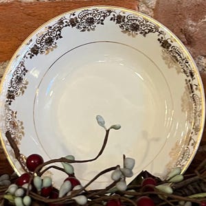 Puede incluir: Un tazón de cerámica blanca con un diseño floral dorado y negro alrededor del borde. El tazón está vacío y tiene una ligera curva en la parte inferior.