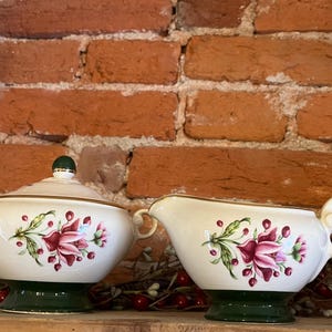 Peut inclure: Un sucrier crème avec un couvercle et une saucière, tous deux avec une base et des poignées vertes. Chaque pièce est décorée d'un motif floral dans les tons de rose, vert et rouge. L'ensemble est placé devant un mur de briques.
