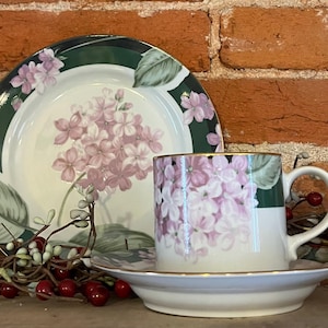 Può includere: Un set di tazza da tè, piattino e piatto bianchi con un motivo floreale. La tazza e il piatto presentano un motivo floreale rosa e bianco con accenti verdi. I bordi sono rifiniti in oro.