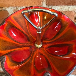 May include: A vibrant, mid-century modern serving dish with a striking red and orange floral design. The dish features a silver handle and a gold-toned central post. The rim has a dark brown edge, adding contrast to the bright colors.