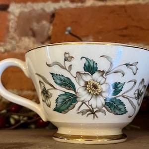 Puede incluir: Taza de té blanca con diseño floral. Presenta una gran flor blanca con centro amarillo y hojas verde azulado. El borde y la base son dorados, con un asa curvada. Probablemente parte de un juego de té.