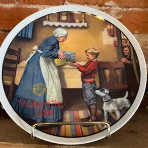 May include: A decorative plate with a Norman Rockwell illustration for Mother's Day 1986. The image shows a woman in a blue dress and apron pouring something into a boy's hands, with a dog and another woman in the background.