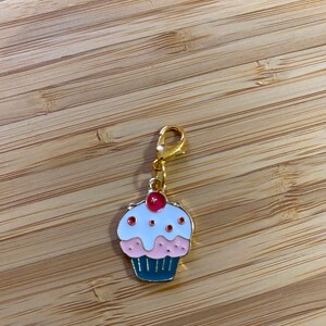 Puede incluir: Un charm de oro en forma de cupcake con glaseado rosa y blanco y una cereza roja en la parte superior. El cupcake está en una cadena de oro.