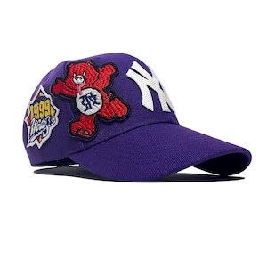 Könnte beinhalten: Violette Baseballkappe mit gebogenem Schirm. Mit einem weißen New York Yankees-Logo, einem roten Bären-Patch und einem Patch mit dem Text "1999 World Series". Der Bären-Patch hat den Text "BR" auf der Brust.