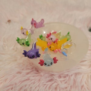 May include: A collection of colorful axolotl figurines in a white bowl. The figurines are made of plastic and have a glossy finish. They are all different colors, including pink, blue, green, yellow, and purple.