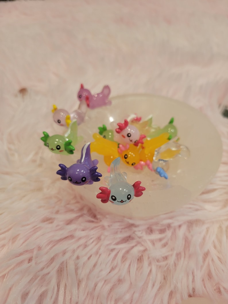 May include: A collection of colorful axolotl figurines in a white bowl. The figurines are made of plastic and have a glossy finish. They are all different colors, including pink, blue, green, yellow, and purple.