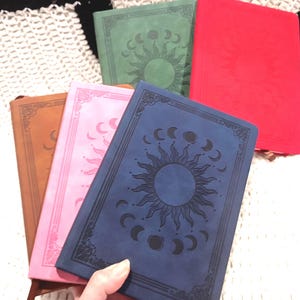 May include: A collection of five rectangular journals in blue, pink, brown, green, and red. Each journal features a debossed cover design showing a central sun surrounded by a lunar cycle phase pattern, enclosed within a decorative rectangular border.