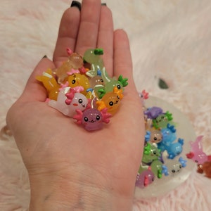 May include: A collection of colorful, small, axolotl figurines with smiling faces. The figurines are made of resin and have a glossy finish.